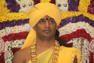Close portrait of a man in a yellow turban and yellow silk garment, with a red tilak mark on his forehead and large gold beaded ceremonial necklaces around his neck. He sits before a backdrop of garlands of yellow marigolds, white and purple flowers, and red roses. The man is the only person visible. Swamiji (probable): long hair pulled back, a yellow turban, saffron-toned clothing and ceremonial gold necklaces, shown in person and seated in a flower-decorated setting.