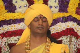 A close photo of a man with eyes closed in meditative stillness, wearing a yellow-saffron turban with a red tilak on his forehead, a yellow shawl draped over bare shoulders, and a heavy gold necklace of large round beads. Behind him is a dense backdrop of yellow, white, purple and red flower garlands with silver-blue decorative accents. Probable Swamiji (appearance cues: yellow turban, gold beaded necklace, yellow shawl) shown in a meditative pose during a satsang setting.