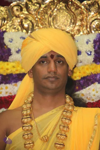 Close-up photo of a man seated before a backdrop of gold ornamental carving and garlands of white, yellow, purple and red flowers. He wears a yellow turban and yellow robe with a gold border, a red bindi on his forehead, and a heavy gold necklace of large ornate beads with a long gold chain. His expression is calm and direct.