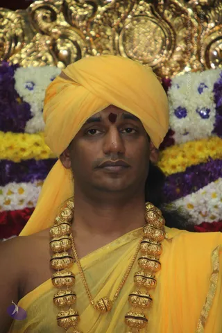 Close-up photograph of a man seated before a backdrop of garlands of white, yellow, purple and red flowers and an ornate gold-toned decorative arch. He wears a bright yellow-orange turban, a matching yellow-orange shawl draped over the shoulder and body, and a heavy multi-strand gold necklace of large ornate beads with a gold chain. A red tilak marks the centre of his forehead. Probable Swamiji: yellow turban, saffron-orange garments and ornate gold necklace, with appearance cues consistent with the page's Swamiji label, but the identification is uncertain.