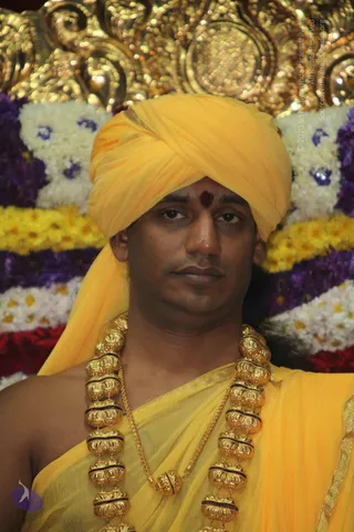 A close-up photograph of one man seated in front of a floral backdrop of white, yellow and purple garlands. He wears a yellow turban and a yellow shawl draped over his shoulders, with a red tilak on his forehead. He has large ornate gold necklaces with rounded beads, and a plain gold chain beneath them. Behind him is an ornate gold-toned decorative throne element.