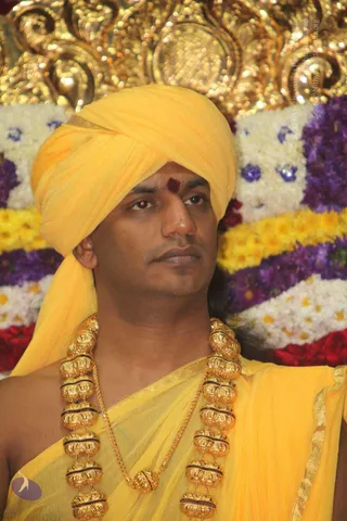 A close-up photograph of a man wearing a yellow turban and yellow draped garment, with a red tilak mark on his forehead and heavy gold beaded necklaces with a thin gold chain. Behind him is a backdrop of white, yellow, purple and red flower garlands and an ornate gold decorative arch. His expression is calm and serious. Swamiji (probable): the man has a turban, saffron-yellow ceremonial clothing and ornate gold necklaces, which are the cues this image offers; the appearance is only a probable match.