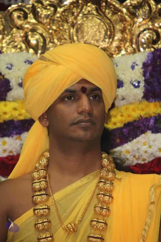 Close photo of a man seated in front of a flower-decorated backdrop of white, yellow and purple blossoms with a gilded ornate panel above. He wears a bright yellow turban and a yellow shawl with a gold border, a red bindi-style mark on his forehead, and a heavy gold ceremonial necklace of large ribbed beads with black bands. Gold-tone chain beads hang at his chest. Appearance cues (yellow turban and shawl, ceremonial gold necklace) are consistent with a person candidate.