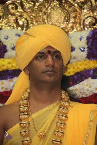 Close-up photograph of a man in a bright yellow turban and matching yellow shawl over the shoulders, with a red tilak mark on his forehead. He wears heavy gold ceremonial beads in a long layered necklace and a thinner gold chain. Behind him is a backdrop of white, yellow, purple and red flower garlands and an ornate gold decorative arch.