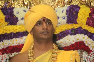 A close-up photograph of a man seated before a backdrop of garlands of yellow marigolds, purple flowers and white blossoms. He wears a bright yellow turban and a yellow shawl draped over the shoulders, with a red kumkum mark on the forehead. He wears a heavy gold ceremonial necklace of large beads over his chest. His gaze is directed to the side with a calm, steady expression. Gold-colored ornamental metalwork is visible at the top edge of the frame.