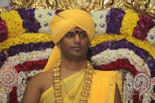 A photograph of a single man seated in front of a floral backdrop of yellow marigolds, purple and white flowers and red roses. He wears a yellow-orange turban with a red tilak mark on his forehead, a yellow-orange shawl draped over one shoulder, and a heavy gold necklace of large beaded bead-like pendants with a long gold chain. Behind him are ornate silver-and-red jewelled decorations. He is seated in a ceremonial setting, appearing as a dignified central figure before flower decorations. Swamiji (probable) cues: a yellow-orange turban and shawl, a red tilak and ornate gold necklace, seated in a ceremonial floral setting. Appearance is consistent with the page's claim but not unmistakable.