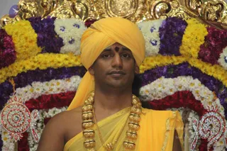 A photograph of a single man seated in front of a floral backdrop of yellow, purple, white and red flowers with a gilded arch. He wears a yellow-orange turban with a red tilak on his forehead, a yellow-orange shawl draped over his shoulders and body, and a heavy ceremonial gold necklace of large beads. Silver-and-red ornamental pieces appear at the lower corners of the frame.