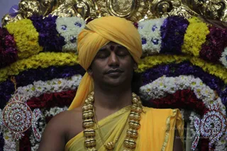 A man seated with eyes closed in meditative stillness, wearing a bright yellow turban and a yellow shawl draped over bare shoulders, with a red tilak mark on his forehead. He wears a heavy gold necklace of large ornate beads with black enamel accents and a thin gold chain. Behind him is an arch of flowers in yellow, purple, white and red marigold and chrysanthemum garlands, with ornamental metal-and-gem decorations at the lower edges. The scene suggests a seated satsang or darshan setting, with the seated figure's appearance matching the page's a person reference only in part, so identity is left uncertain.