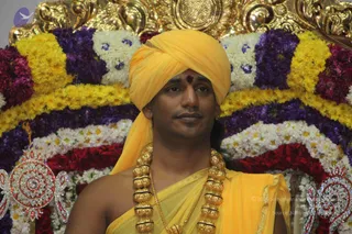 A single man sits in front of a floral backdrop of yellow, purple, white and red flower garlands arranged in tiers, with an ornate gold and gem-studded frame above. He wears a bright yellow turban, a yellow shawl draped over his bare shoulder, and a thick layered gold necklace; a red tilak marks his forehead. Decorative jewelled plates flank the lower corners.