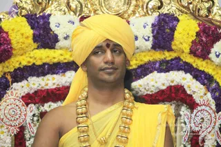 Close photo of a man seated in front of a dense floral backdrop of yellow marigolds, purple flowers, white blossoms and red roses. He wears a bright yellow turban with a small red tilak on the forehead, a yellow shawl draped over his bare shoulder and yellow dhoti-style cloth, and a heavy necklace of large gold beads and a gold pendant. Ornate gold-and-red jewelled floral motifs flank the arrangement at the lower corners.