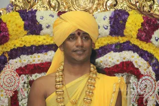 A man stands centrally in front of a floral backdrop of marigolds, purple flowers and white blossoms arranged in arching rows, with red roses below. He wears a yellow turban with a small red tilak on his forehead, a yellow shawl draped over bare shoulders, and a heavy ceremonial gold necklace of large beads. Ornate gold and jewelled decorations flank the arch. Swamiji (probable) appears in person, shown by the yellow turban, gold beaded necklace and ceremonial yellow garments, seated before the floral display during a satsang.