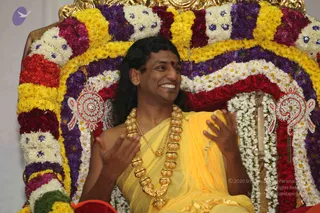A man with long dark hair and a red bindi sits smiling on a throne decorated with garlands of yellow, purple, red and white flowers, with blue-jewelled flower accents. He wears a yellow silk garment over bare shoulders and a heavy gold necklace of large bead-like pieces, with a thinner gold chain beneath. His hands are raised in an animated gesture as he speaks. Ornate silver-and-red decorative panels hang on either side of the throne back.