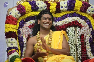 A man with long dark hair and a small red bindi sits on a throne decorated with bands of yellow, purple, red and white flower garlands and silver-and-red ornamental plaques. He wears a yellow-orange cloth draped over his bare shoulders and a heavy gold multi-bead necklace, and he smiles broadly while raising his hands in an expressive gesture during a satsang. The setting is a flower-decorated seat with a white backdrop. The appearance matches a person (long swept hair, ornate gold necklace, saffron-yellow clothing, throne) seated in a satsang setting.