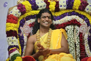 A photograph of a smiling man seated on a throne decorated with a dense arch of flower garlands in yellow, purple, red and white, with blue-centred white blossoms. He wears a saffron-yellow shawl draped over his bare shoulder and a heavy multi-strand gold necklace, with a red-and-gold bindi on his forehead and long dark hair. His hands are pressed together in front of his chest in a clapping or greeting gesture. Ornate gold-and-silver ceremonial emblems hang on either side of the throne arch. A red cushion is visible in the lower foreground. Swamiji (probable) is present as a seated central figure, shown in person with his long hair, saffron shawl and gold necklace.