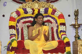 A man seated cross-legged on a red-cushioned throne framed by an arch of marigolds and white flowers in yellow, purple, red and white. He wears a yellow silk garment, a heavy gold necklace of beads, and a small red bindi, and is smiling while clapping his hands. A brass oil lamp with a lit flame stands at right beside a golden-trimmed stand, and a red and white decorated backdrop hangs behind. The swami-like presentation with long swept-back hair and gold ornaments is consistent with a person seated in honour at a satsang, and the lamp and garland setting marks a formal discourse.