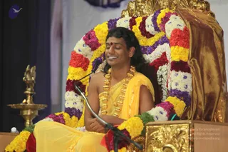 A photograph of a seated man at a satsang on a stage, on a throne-like gold chair backed by a large arc of marigold, rose-coloured and white flower garlands. He wears a yellow dhoti and matching yellow shawl over a bare chest, with a heavy gold multi-bead necklace and a red bindi, and long dark hair swept back. He sits cross-legged with his hands folded, smiling, in front of a microphone stand. A brass oil lamp with a lit flame stands to the left, and a gold-draped backdrop fills the right side.