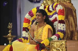 A seated man with long, swept-back dark hair and a red bindi wears a yellow dhoti-style garment with a yellow shawl and a thick gold ceremonial necklace of beads. He sits on a golden throne decorated with a dense arc of yellow, red, purple and white flower garlands, with a microphone in front of him. A brass oil lamp with a lit flame stands at left, and a gold-toned ornamental panel is at the lower right. Appearance cues suggest a person, seated on a peetha and speaking at a satsang.