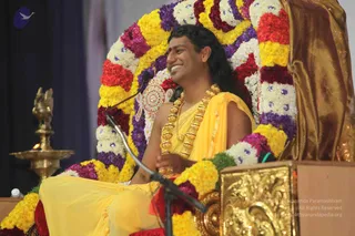 A man with long dark hair sits smiling on a throne-like seat decorated with garlands of yellow marigolds, white flowers and purple blooms. He wears a yellow dhoti and wrap, a gold rudraksha mala necklace, and a gold armlet, with his hands folded at his waist. A microphone stand is in front of him, a brass oil lamp with a lit flame stands at the left, and a gold-draped backdrop fills the right side. Probable Swamiji (long swept-back hair, ornate gold necklaces, saffron-yellow clothing, seated on a garlanded throne), seated in satsang. Only one person is visible.