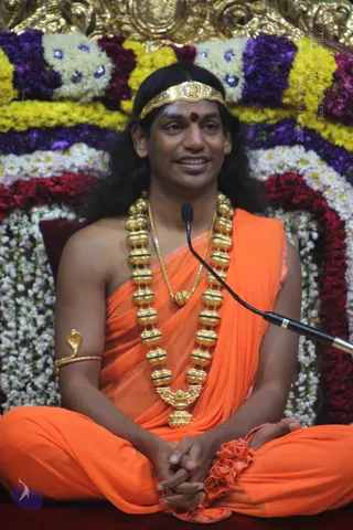 A seated man in saffron-orange robes, bare-shouldered, speaks into a gooseneck microphone in front of a floral backdrop of marigold, white, purple and red garlands. He wears a gold headband with a red bindi, long swept-back dark hair, a heavy gold multi-bead garland with a central ornament, a gold chain, and a gold cobra armlet. His hands rest clasped in his lap in a cross-legged posture. Swamiji (probable): the saffron robes, long swept-back hair, gold headband and ceremonial gold necklaces are consistent with the page's identification of the speaker as the satsang discourse presenter.