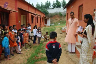 A photograph of an outdoor courtyard between terracotta-red brick buildings with corrugated awnings, with a dirt path and patches of grass. A man in a peach-coloured long kurta and white dhoti, with a rudraksha mala around his neck, stands on a step facing a group of children. To the right, a woman in a cream sari with a gold border holds a clear plastic container. Around thirty children, some barefoot and wearing orange, blue and navy clothes, gather on the left, while a young boy in a navy and red sweatshirt with the number 5 stands with his back to the camera in the foreground. A person holding a camera is partly visible at the upper right.