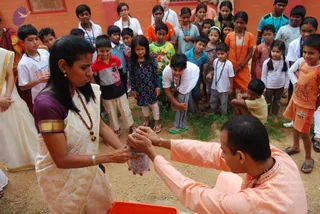 An outdoor photograph on bare dirt ground beside a terracotta-coloured wall, showing a woman in a cream silk sari with a purple blouse and gold border and a rudraksha-style beaded necklace, leaning forward to take a small clear bottle from a man in a peach-coloured kurta with a bangle on his wrist, who holds the bottle over an orange plastic tub. A crowd of children in mixed casual clothes, several wearing lanyards and white shirts, stands and watches closely, with a boy in an orange shirt at the right edge.