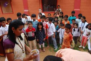 A woman in a cream sari with a purple border and rudraksha mala stands at left holding a clear plastic container of food, possibly prasadam or annadan, while a crowd of children gathers around her in a dirt courtyard beside an orange-red wall with a barred window. Many children wear white t-shirts, lanyards and rudraksha-style beads; a boy in a red striped shirt and a boy in an orange printed shirt stand near the centre. At the bottom right a person in a peach-coloured garment bends forward toward the crowd, partly out of frame.