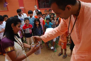 Outdoor photo at an orange-walled building with a dirt ground. A man in a peach-orange kurta with rudraksha mala beads bends forward and reaches into a clear plastic jar of grains or prasadam held by a woman in a cream sari with a purple blouse, gold border and red bead necklace. Around them a group of about fifteen children and a few teenagers stand watching and smiling, some in colourful shirts and one in a white t-shirt, with a barred window behind. The scene suggests food being distributed to children, an annadan style giving. A purple circular logo appears in the upper left corner.