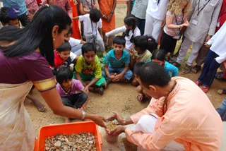 A photograph of a community gathering on a dirt ground with many children seated and standing around. In the foreground a woman in a purple blouse and cream sari with gold border holds out her hands over an orange plastic tub of small stones. Beside her a man in a peach-orange kurta with a rudraksha mala and white dhoti kneels and holds a small cup of granules. Children in bright shirts and shorts watch closely, and adults in white and striped clothing stand at the back, some wearing lanyards.