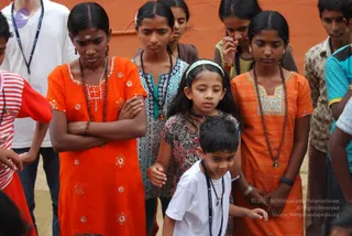 A group of children and teenagers stand closely together outdoors against an orange-red wall, looking downward with serious expressions. A girl in front wears an orange embroidered kurta with sequined details, a rudraksha mala, and a vermilion bindi, with her arms folded. Beside her a girl in a blue-and-white patterned top wears a dark rudraksha mala, and a younger girl in a floral dress with a white headband stands in the middle. Another girl on the right wears an orange kurta with a rudraksha mala and a pendant, and a boy in white with a lanyard stands in the foreground. A man in a white T-shirt with a lanyard stands at the upper left. No a person candidate is visible in the scene.