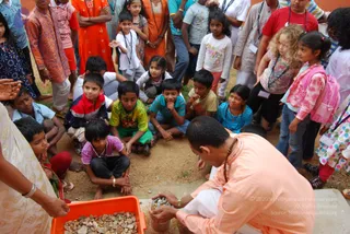 A crowd of about twenty children and teens gathers in a dusty outdoor yard around a man crouched on a concrete edge. He wears a peach-orange kurta with a rudraksha-style beaded necklace and white dhoti, and is handling small stones from a cup while an orange plastic tray of gravel sits in front. Children sit cross-legged and stand around him, several wearing lanyards with badges. A woman in a cream sari with gold bangles stands at the left edge, and a girl in pink jacket and backpack stands at right.