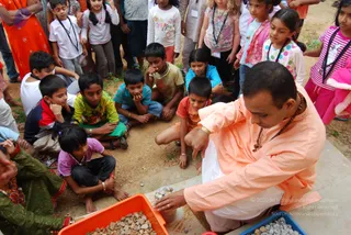 A photograph of a man in a peach-orange kurta over white dhoti, with a rudraksha mala around his neck and a gold bangle, kneeling on a concrete slab and sorting small pebbles from a bowl into an orange plastic tray. A semicircle of children sits and stands around him, watching intently; a girl in pink and polka dots, a boy in a yellow-green shirt, and a girl in a blue top are prominent, and a woman in green salwar kameez with a sari-style wrap stands at the left edge. Blue and orange tubs of gravel sit at the front. Children wear lanyards with name badges. Dusty ground and greenery form the background.