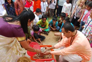 A photograph of a ground-level ritual at an outdoor site. A seated man in a peach-orange kurta and white dhoti, with a rudraksha mala and bangle, holds a small silver dish of stones over a brown clay pot set in an orange basket of gravel. A woman in a purple blouse and cream sari with gold border, also with bangles, pours from a red bowl beside him. Around them, about a dozen children and a few adults stand and crouch, watching closely, several wearing name badges. Gravel and sand lie on the ground.