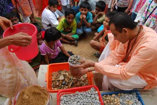 A photograph of a man in a peach-orange kurta with a beaded rudraksha-style mala and white dhoti crouches at a concrete ledge, holding a clear cup of small grey and brown stones toward the viewer. Orange and red plastic tubs hold heaps of gravel and crushed stone, and a blue tub holds more stones. At the left edge, a woman in a pale pink sari with gold border pours from a red bucket. Behind them, a group of about a dozen children sit and stand on dirt ground, watching closely; one girl in a lavender top sits in the centre. Some children wear lanyards.