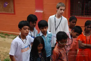 A group of children and one older teenager stand outdoors in a sandy courtyard before a red-and-orange painted wall with a barred window. A tall teenage boy in a white t-shirt with a lanyard stands at the back, and a boy in a white t-shirt printed with a spelling bee logo stands at the left. Several children wear rudraksha-style beaded necklaces; a girl in an orange dress with beaded jewellery stands at right. Most of them look downward with eyes closed or lowered. No a person candidate is visible.