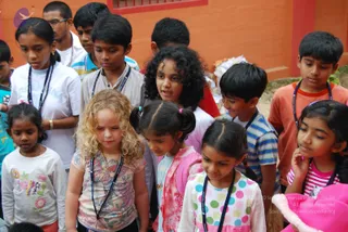 A photograph of a group of children and a few teenagers standing closely together outdoors in front of a bright orange wall with a small barred window. A young woman in a white t-shirt with a printed logo stands at left, and a boy in a striped shirt has a small red mark on his forehead. Several children wear dark beaded lanyards around their necks. A curly-haired blonde girl in a floral-print top stands at front centre, and other girls in pink, polka-dot and striped clothes look down with their hands near their faces. No deity, ritual or a person appears in the scene.