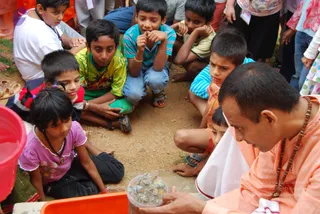 Outdoor photo of a man in saffron-orange robes and a rudraksha mala seated on bare earth, leaning toward a clear plastic container of small shells held in his hands. Around him a group of about a dozen children sit or crouch on the ground, watching intently; one girl in a purple top with a pendant rests her chin on her hands. A red bucket sits at left and an orange tray at lower left. The man's face is turned down and to the side, and his appearance is not a clear, multi-cue match for the named presenter, so no confident identification is made; he is described by role as a man in saffron robes demonstrating to children.