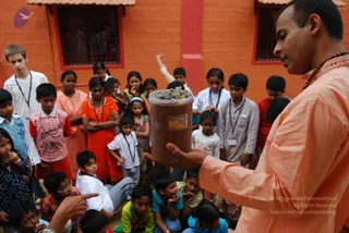 A man in a light orange-peach kurta with a rudraksha mala around his neck holds up a clear plastic jar filled with dark earthen contents and small objects, showing it to an attentive group. Behind him, a crowd of children and young people sits and stands in front of a red-orange brick building with barred windows. Several children wear white shirts with blue lanyard badges, a girl wears an orange salwar kameez, and some are in striped shirts and bright patterned clothes. One child in the foreground points at the jar. The scene looks like a teaching demonstration in a courtyard.