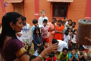 A woman in a plum-coloured sari with gold border stands at left, gesturing with one hand toward a group of children gathered in an outdoor courtyard with red-painted brick walls and a barred window. Many children stand and sit on bare earth, some in school uniforms with white shirts and lanyards, others in colourful clothes such as an orange kurta and pink salwar. At right, a hand holds a cylindrical container filled with dark granular material, possibly herbs or seeds. The scene suggests a group teaching or activity session for young participants. No person is identifiable as a person here.