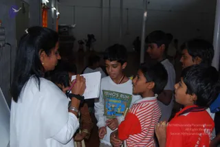 A woman in white clothes with her hair in a bun stands at left holding a black microphone in one hand and a notebook with a pink pen in the other, speaking to a group of boys. Among them, a boy in a white t-shirt with a red and gold lanyard holds a green book titled Primo's Run, while a boy in a red and white striped shirt listens closely. Other boys in grey and red shirts crowd in on the right. In the background, a hall with white pillars and orange drapes shows several more people moving about. No a person candidate is visible in the frame.
