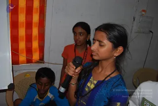 A young girl in a blue silk-bordered salwar kameez with a dark beaded mala necklace stands in the foreground holding a black handheld microphone, eyes closed as she speaks or sings. Behind her, a second girl in an orange top with a red beaded necklace stands by a red and yellow checked curtain. At lower left, a boy in a blue-and-white sports shirt sits on a beige plastic chair. A white wall with an electrical socket and a small figurine on a shelf forms the background, and a partially visible person in white sits at right. The setting suggests a satsang or group programme.