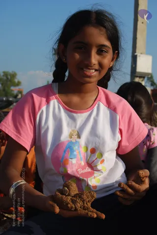 A smiling girl with long dark hair in a single side braid sits outdoors under a clear blue sky, holding a small clay figure of an animal in both hands. She wears a pink and white raglan T-shirt printed with a Barbie graphic of a doll in a blue top above a pink circle with coloured dots. Behind her a concrete utility pole and a blurred second girl in pink are visible. No other people are clearly visible.