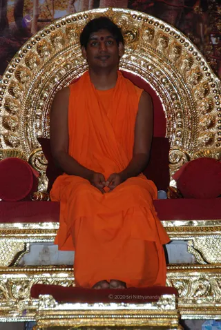 A man in a loose orange saffron robe sits cross-legged on an ornate gold throne with a red cushioned seat and a gilt arched backrest. He has a red dot on his forehead, hands folded in his lap, and a small microphone clipped near the throne. Behind him is a painted forest backdrop. The throne sits on a gold-trimmed platform with a red footrest.
