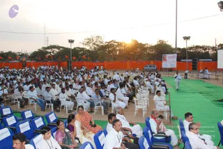 Outdoor evening satsang at a large open ground with rows of white plastic chairs filled by a dense audience, many men in white dhotis and shirts, and some women in saris. Front-row blue chairs are labelled RESERVED. A green mat runs toward a stage, and a large banner with a portrait is set at the right. Orange cloth lines the back fence, orange flags fly, and floodlights stand over the crowd as the sun sets behind trees. A man in white walks across the green mat near the stage. A banner with a portrait labelled Paramahamsa Nithyananda is visible at the right, so no physically present candidate is identifiable in the scene.