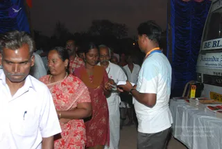 Photograph of an outdoor evening event at dusk. A line of devotees queues beside a table of books and pamphlets draped in white cloth, next to a white bus with blue-curtained canopy. A man in a white t-shirt with a saffron-orange lanyard, glasses and a wristwatch hands a small card or booklet to a woman in a red-and-white floral saree with a red bindi and gold bangles. Beside her a woman in a rust-red patterned salwar kameez with a brown dupatta holds papers, and an older man in white shirt and white dhoti stands with a grey moustache. A man in a white shirt stands at left, and others are visible in the background under the blue drapes.