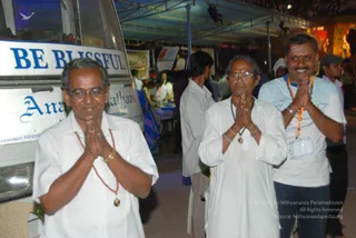 Three men stand in a row outdoors at night beside a white van with the words "BE BLISSFUL" and partial lettering reading "Ana..." and "Nithyanandapuri Kallugopalam" along its side. Each presses palms together in anjali namaste. The man on the left wears a white shirt, a red-orange beaded necklace and a pendant, and gold bracelets. The man in the middle wears white kurta-style clothing, a rudraksha-style mala with a pendant, a vermilion mark on his forehead and glasses. The man on the right wears a light blue t-shirt with orange lanyard and an ID badge, and a bracelet. Behind them a covered open-air hall has blue canopy supports, people walking, a table, banners and a lit decorated backdrop with deity murti figures.