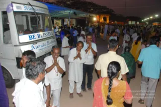 Evening outdoor scene at an event ground with many people walking across a sandy path past a white van with blue lettering reading "Nithyanandam" and "BE BLISSFUL" and "Ananda Rathum Nithyananda Dhyanapeetam". Several men in white dhotis and shirts stand with palms pressed together in namaste greeting. One man in a white shirt wears a long orange-and-brown beaded necklace and a lanyard, another in a white shirt and dark trousers also greets with folded hands, and a man wearing a white turban stands behind them. A woman in a red and orange sari with a long braid is seen from behind in the foreground. Illuminated stalls with banners, a lit temple gopuram-style structure and street lamps are visible in the background.