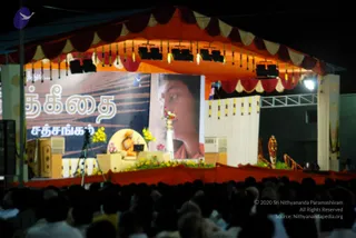 A night-time outdoor satsang stage under an orange and red canopy with white and red valances and hanging gold tassels. A seated figure in orange clothing sits on a yellow cushioned throne surrounded by yellow and pink flower arrangements, with a laptop and a camera on a tripod nearby. A large portrait of a man in orange and pink robes is displayed beside the stage, and a long Tamil-script banner hangs at left. A dense crowd of silhouetted people fills the foreground.