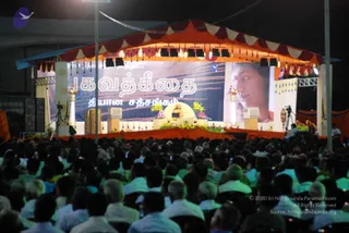Night photo of an outdoor satsang stage under an orange and maroon tented canopy with hanging tassels and spotlights. A seated teacher in orange robes sits on a raised dais decorated with flowers and a central floral arrangement, speaking into a microphone. A large backdrop banner reads Bhagavad Gita and a second line in Tamil script; a large portrait of a man with short dark hair in profile hangs at the right. A dense crowd of several hundred seated people, many in white shirts, fills the foreground. Camera tripods and speakers flank the stage.