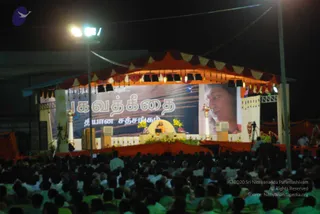 Night photograph of an open-air outdoor satsang stage under an orange and maroon canopy with strings of lights and yellow marigold garlands. A seated figure in saffron-orange clothing sits on a raised throne-like seat at centre stage, with a laptop-style screen and floral decorations nearby. Behind the stage a large banner shows Tamil script and a close-up portrait of a face on a large screen to the right. Speakers and a camera on a tripod flank the stage. A dense crowd of many people in light and white clothing fills the foreground seating area under floodlights on tall poles.