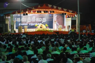 Night-time outdoor satsang with a large crowd of seated devotees filling the foreground, many in white and light green clothing. An orange and maroon canopy with tassels and bunting covers a stage decorated with yellow and pink flowers and white pillars. A figure in orange robes sits on a raised seat at the centre of the stage, with two speakers flanking the platform. A large screen shows a Tamil banner reading 'Bhagavad Gita' and 'Dhyana Satsangam', plus a close-up portrait of a woman. Camera tripods and stage lights are visible at the sides.